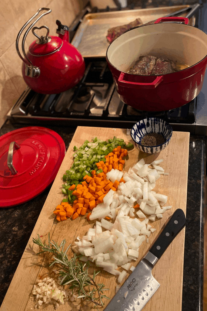 chopped mirepoix on cutting board next to red dutch oven with short ribs inside