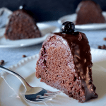 slice of chocolate cake on white plate next to fork with more cake in background