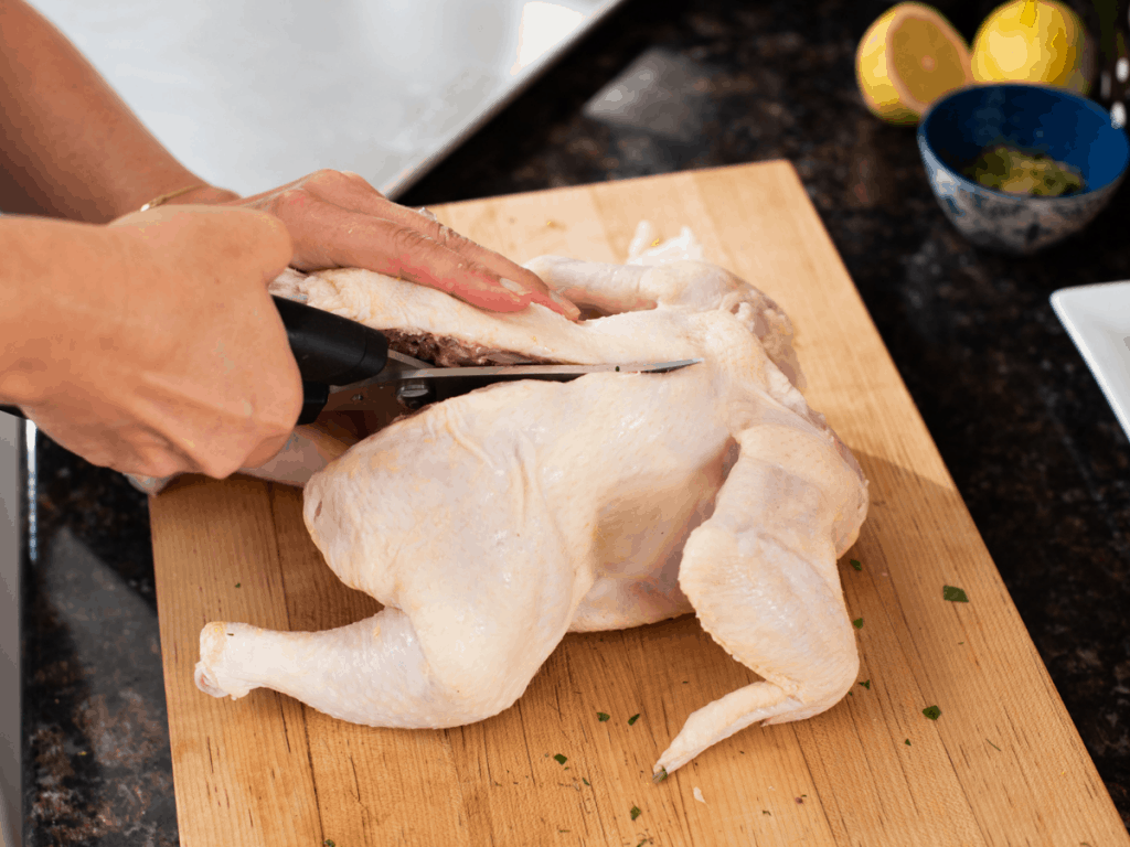 woman's hands using kitchen sheers to cut backbone from raw chicken on wooden cutting board