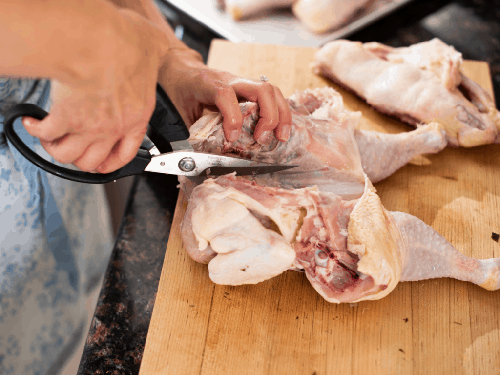 womans hands snipping breastbone of raw chicken with kitchen sheers on wood cutting board