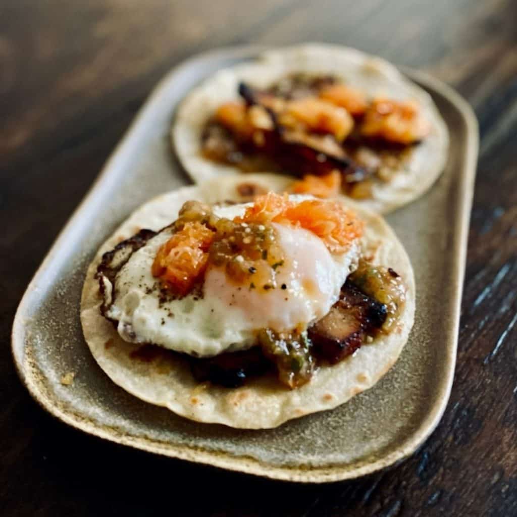 two corn tortillas with pork belly, salsa and fried eggs on square plate