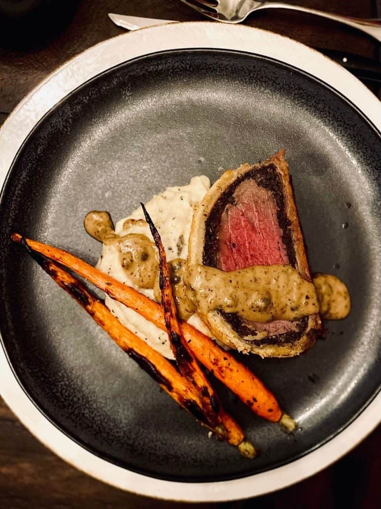 Slice of rare beef wellington with potatoes and carrots and sauce on black plate