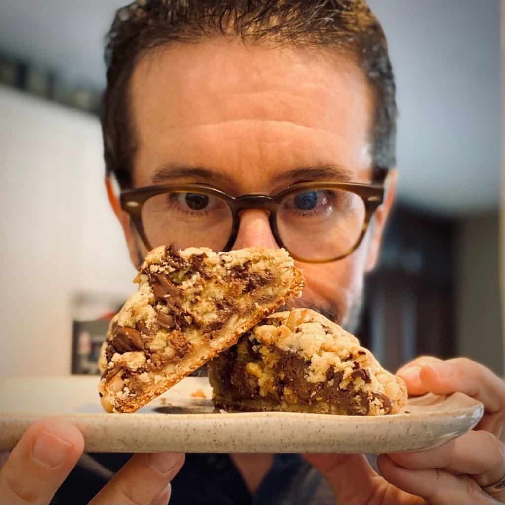man with glasses looking at plate with huge chocolate chip cookie cut in half