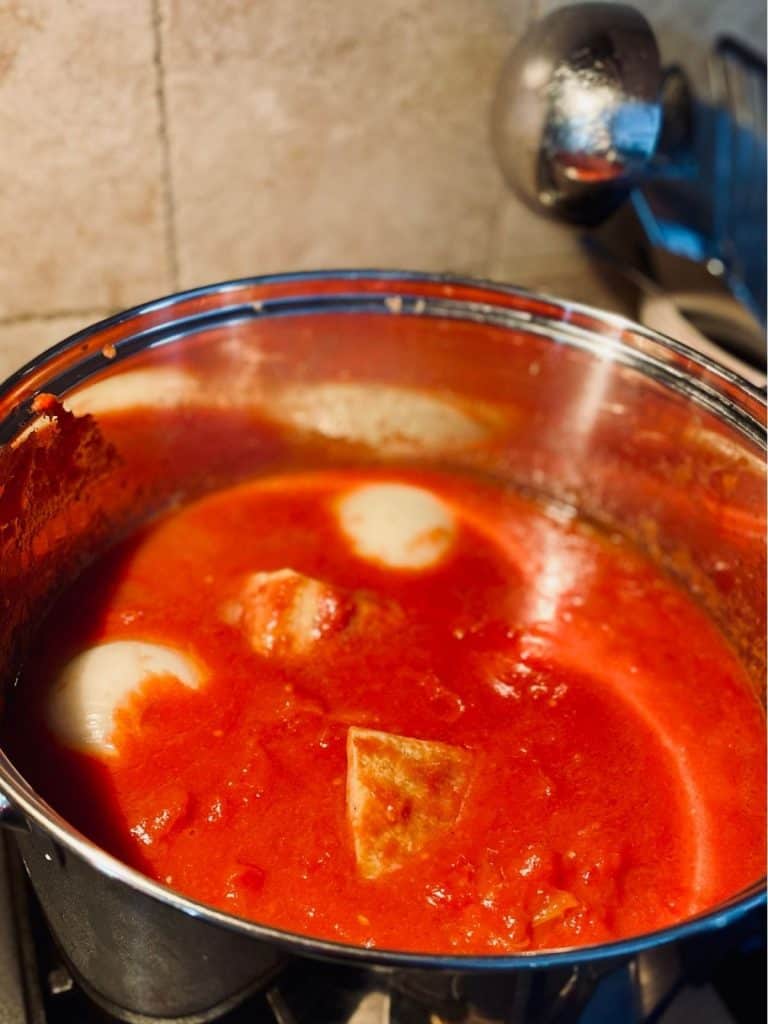 stockpot of simmering marinara sauce with onion halves and parmesan cheese rinds