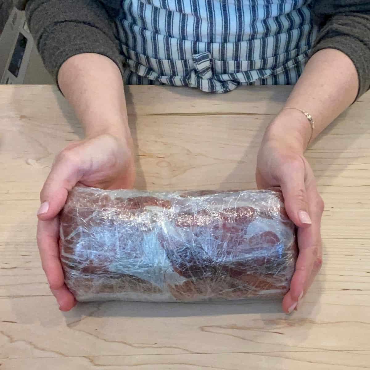 two hands holding a prociutto wrapped tenderloin wrapped in plastic