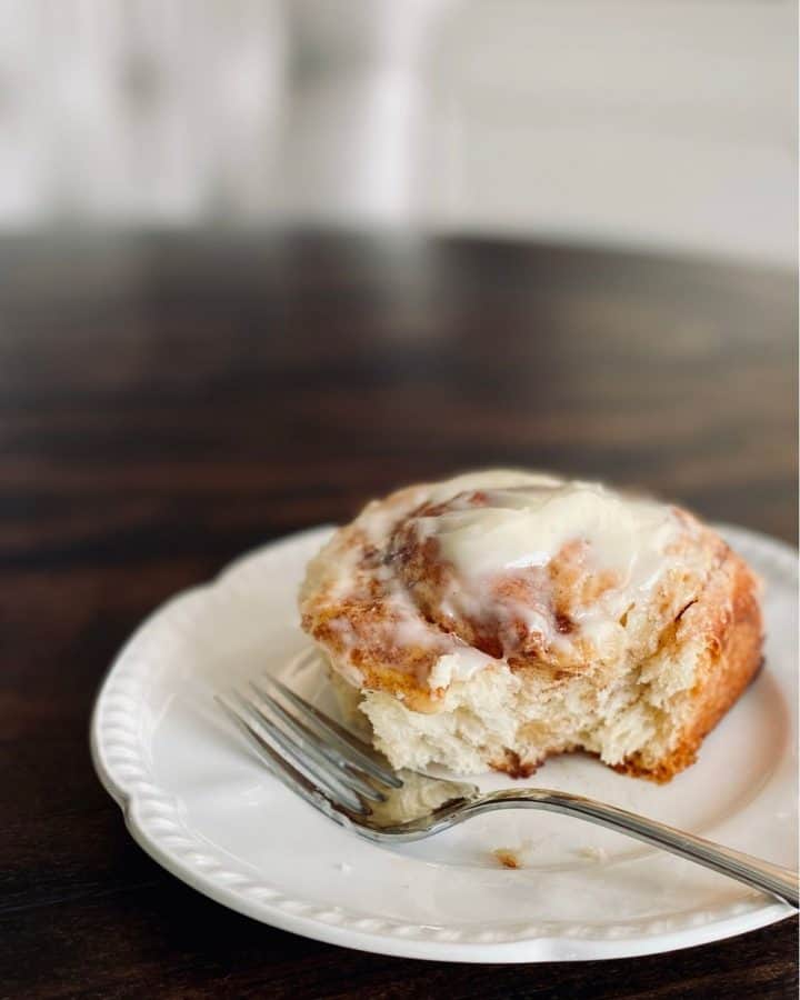 iced cinnamon roll on white plate next to fork with bite taken out
