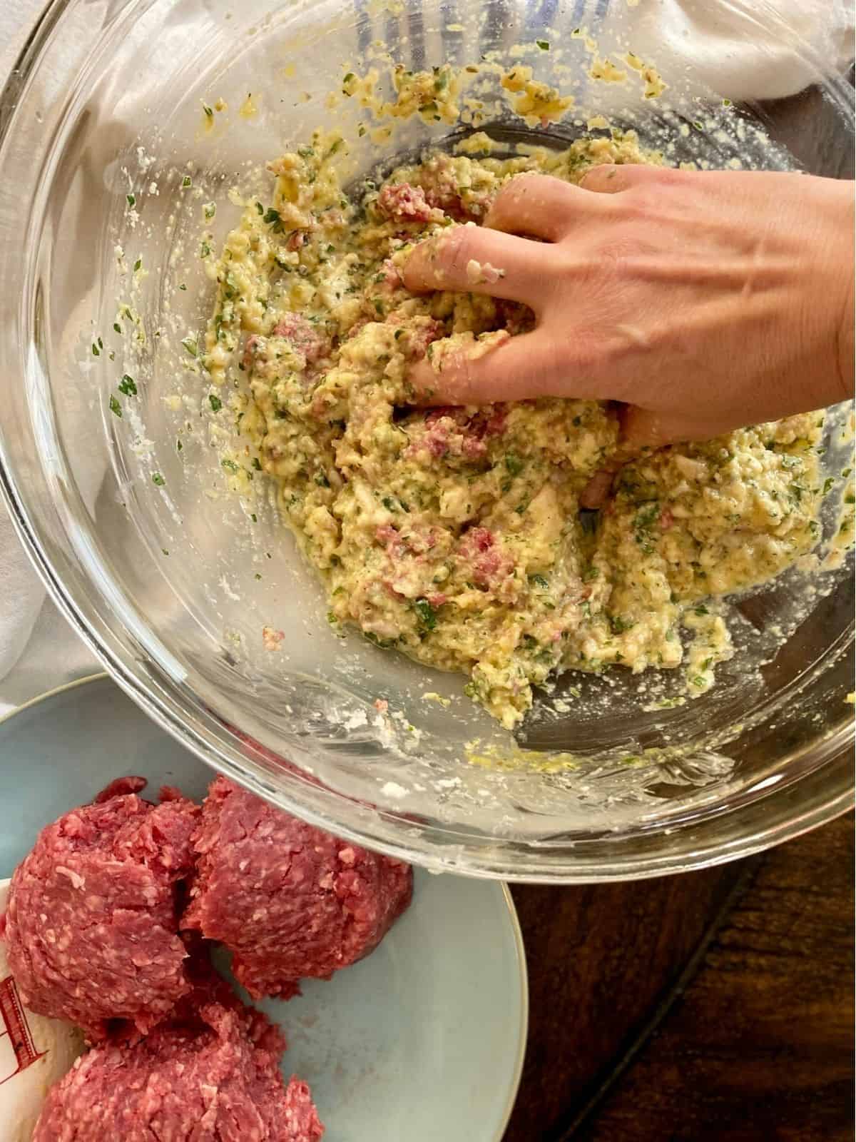 hand mixing meatball ingredients in glass bowl next to plate of ground beef