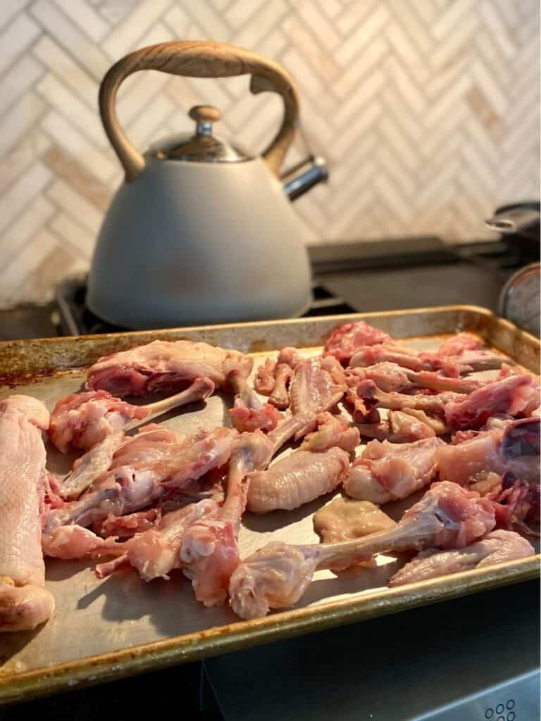 baking sheet filled with raw chicken bones and parts on stove top with tea kettle in background