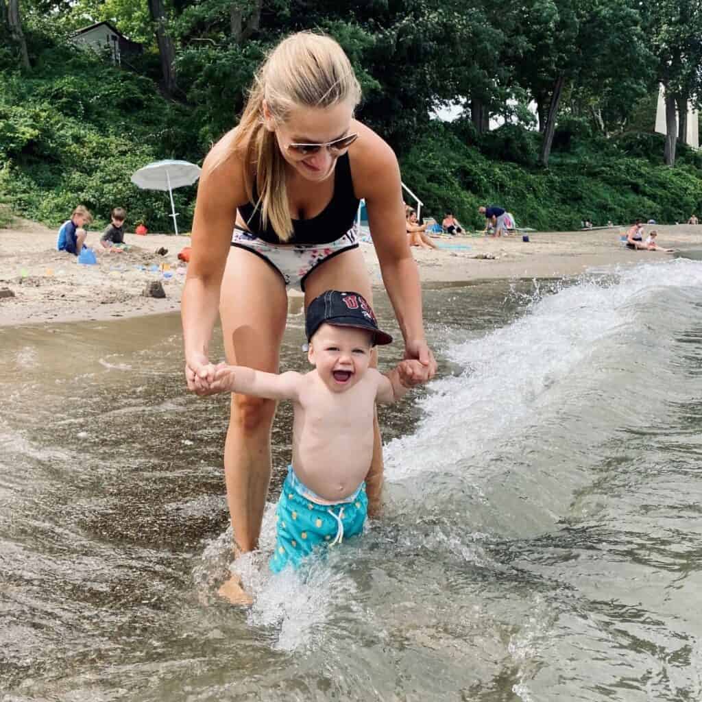 baby holding moms hands as he's splashed by waves