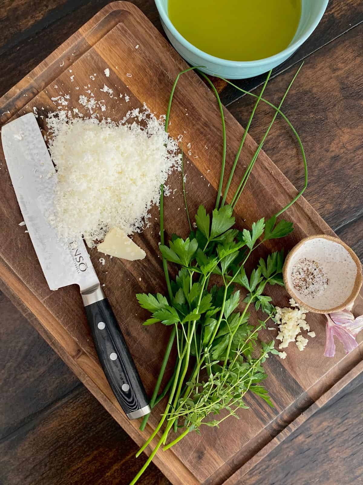bowl of olive oil on wood board next to knife, grated parmesan and fresh herbs