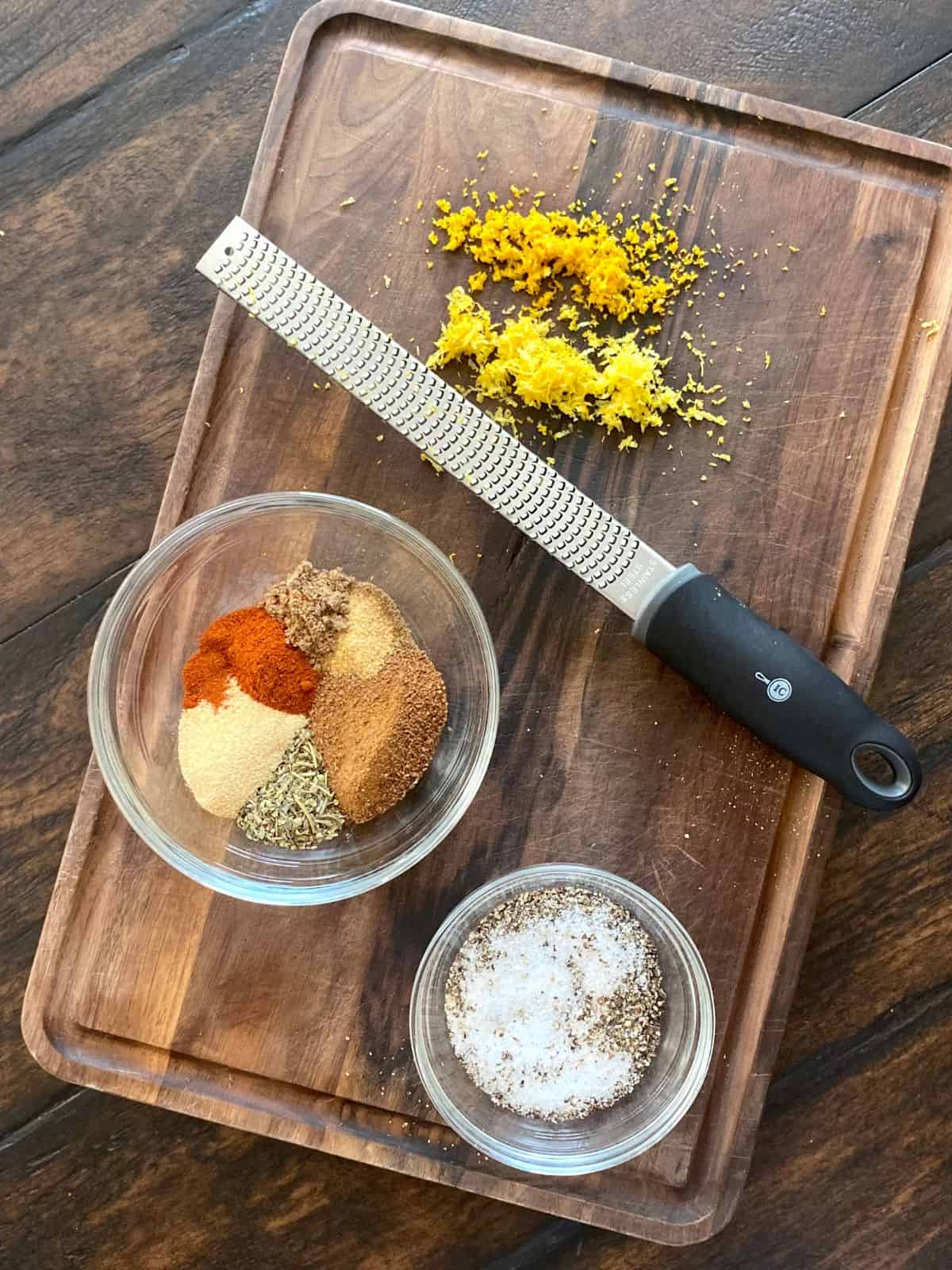 spices, zest, salt and pepper in bowls on wood board next to microplane