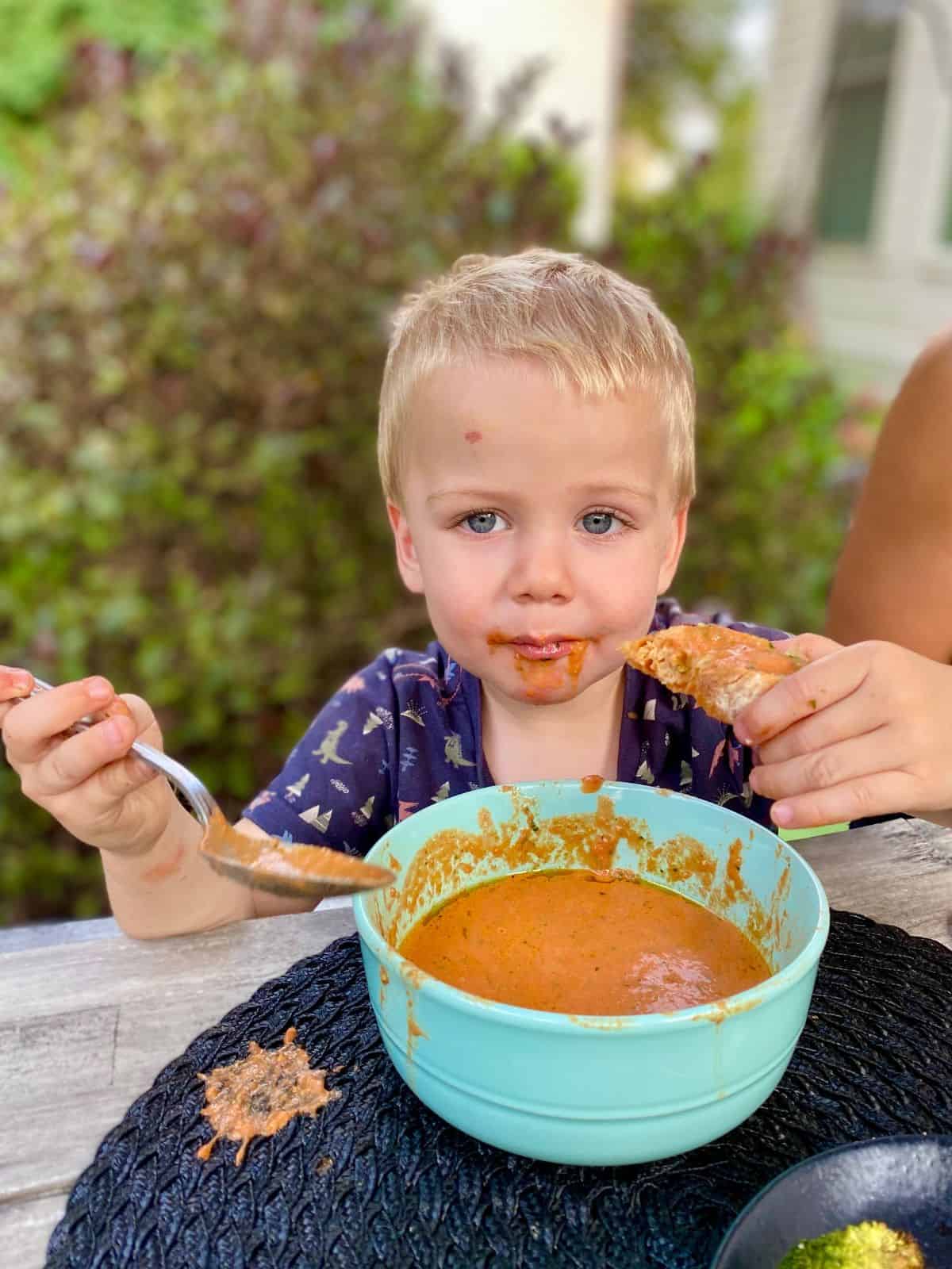 little boy eating tomato soup and holding a soup-dipped crostini
