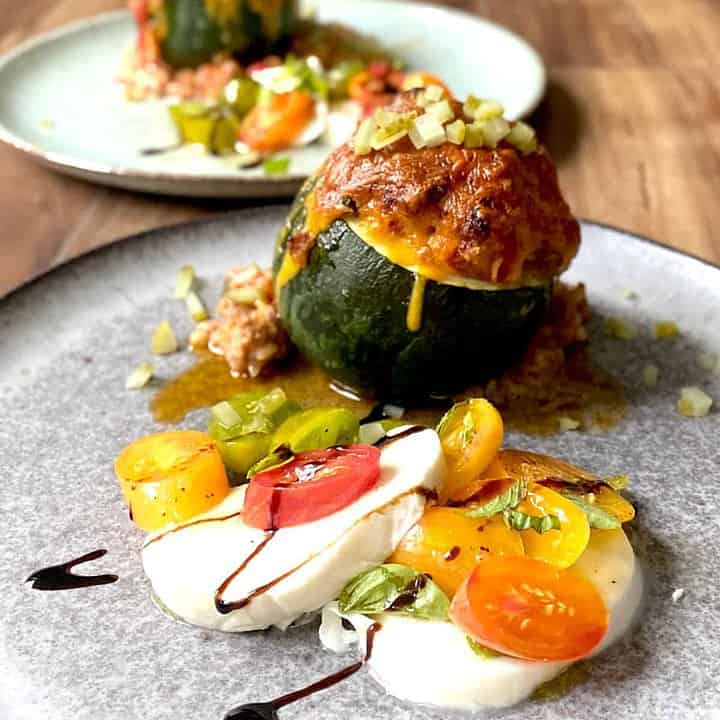 cheeseburger stuffed zucchini and caprese salad on grey plate with another plate of food in background